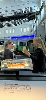 Zwei Personen betrachten ein illuminiertes Modell einer Bahnanlage im SüdBahn Museum Mürzzuschlag. Im Hintergrund eine große, lichtdurchflutete Halle.